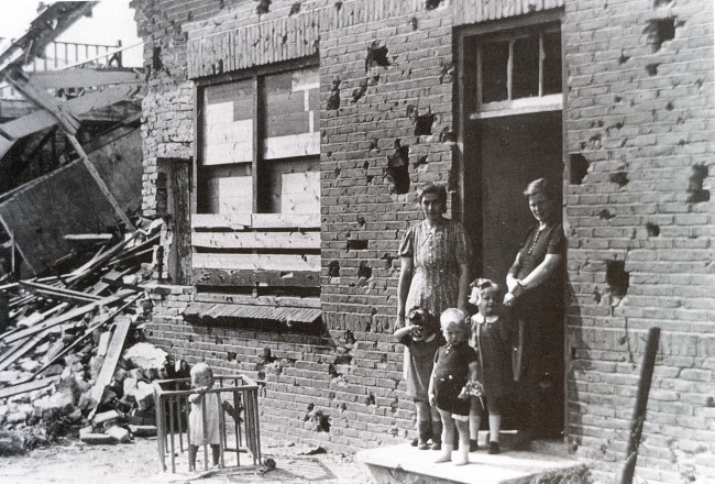 In de deuropening van een verwoest huis staan twee vrouwen en drie kinderen. Links voor het huis een kindje in een babybox.