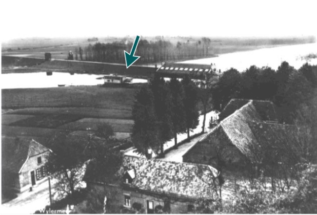 There are houses with trees by a road in the foreground. There is a river with a lock in the background.