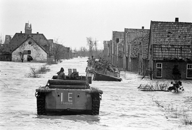 Überschwemmte Siedlung mit Häusern. Im Wasser fahren Amphibienfahrzeuge auf denen Personen sitzen.