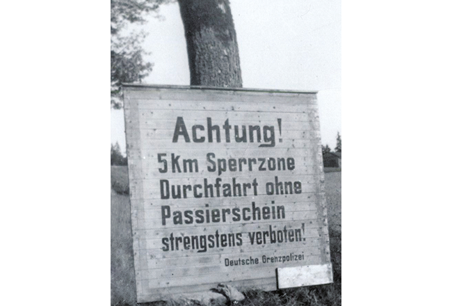 Holzschild an einen Baum gelehnt mit der Aufschrift: „Achtung! 5 Km Sperrzone Durchfahrt ohne Passierschein strengstens verboten! Deutsche Grenzpolizei“.